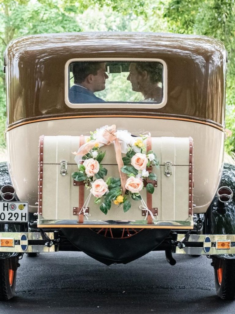 Das Braupar auf dem Weg zur Trauung Brautpaar sitzt im Brauttaxi mit Blumenschmuck am Kofferraum.